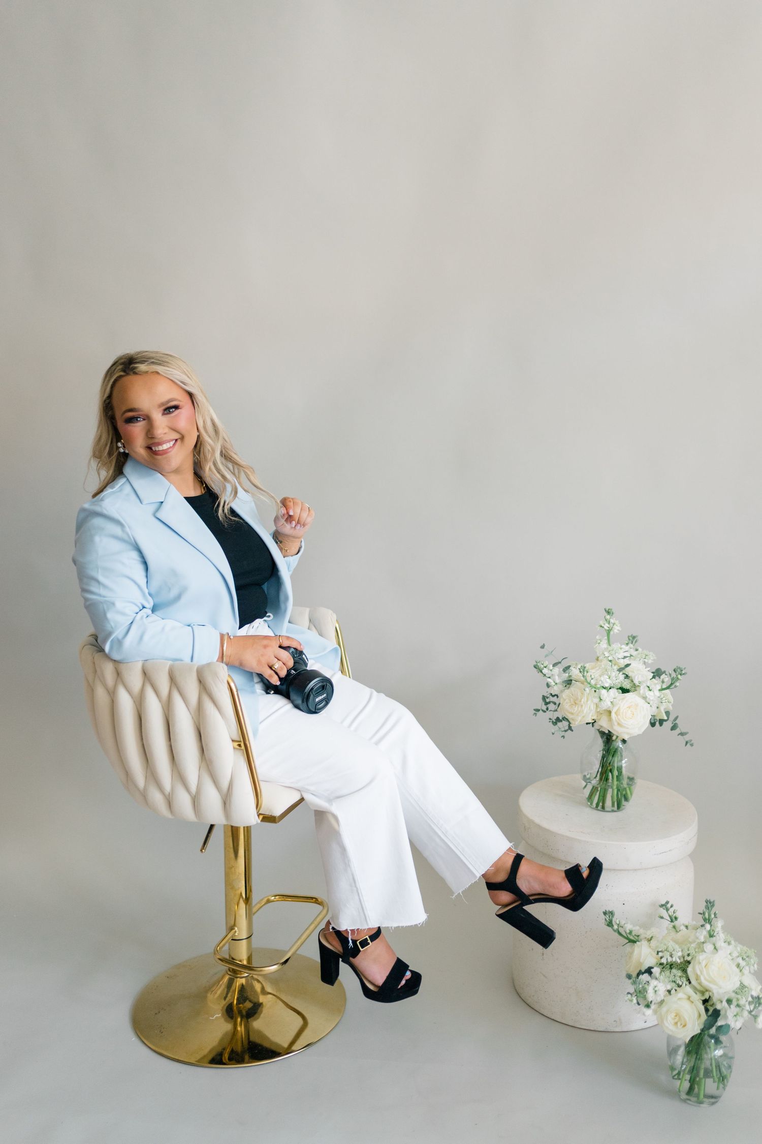 Smiling blonde woman in light blue blazer and white pants sits in gold chair holding camera beside white floral arrangement.