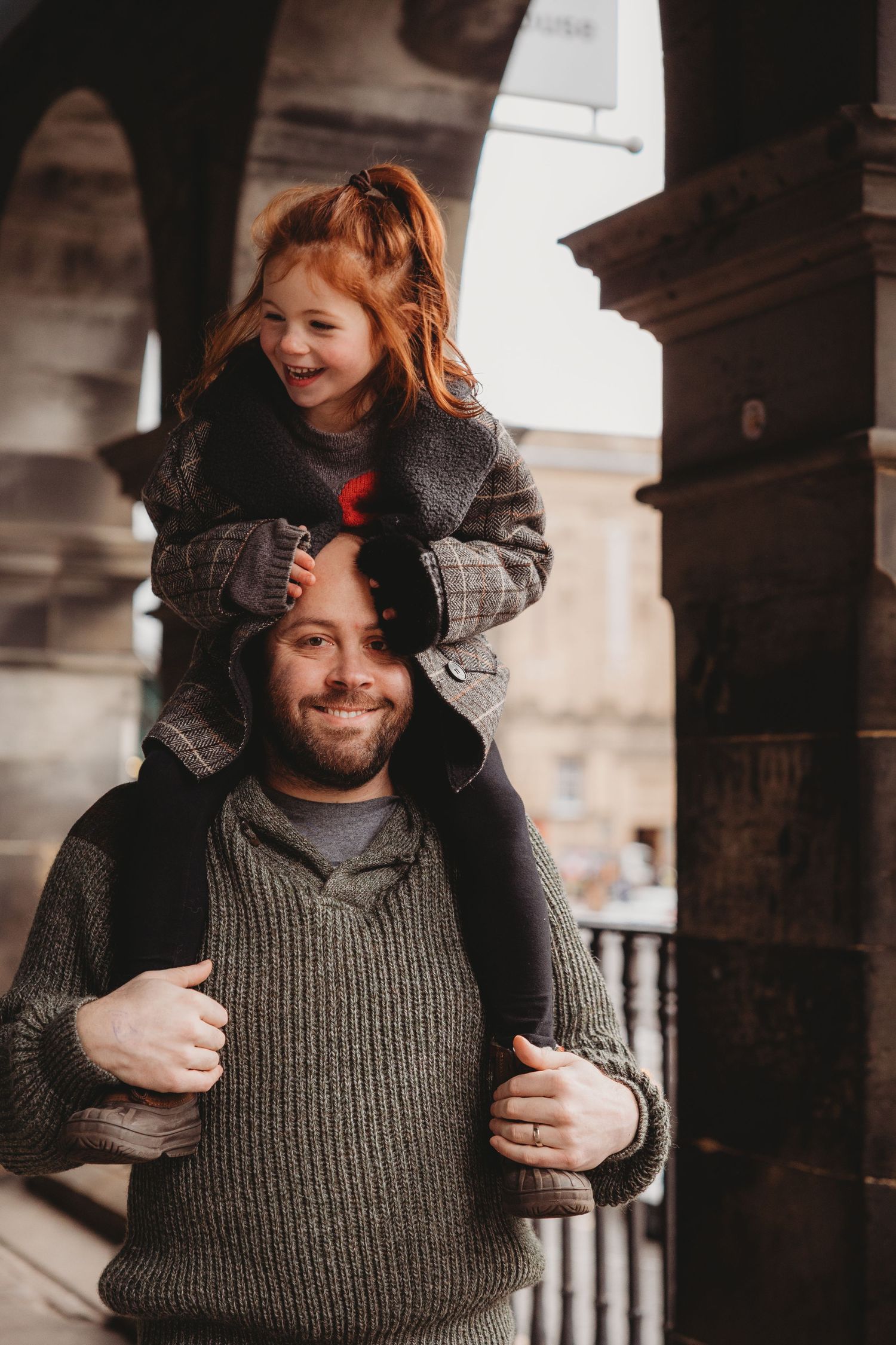 A smiling man carries a laughing red-haired girl on his shoulders under a stone archway.