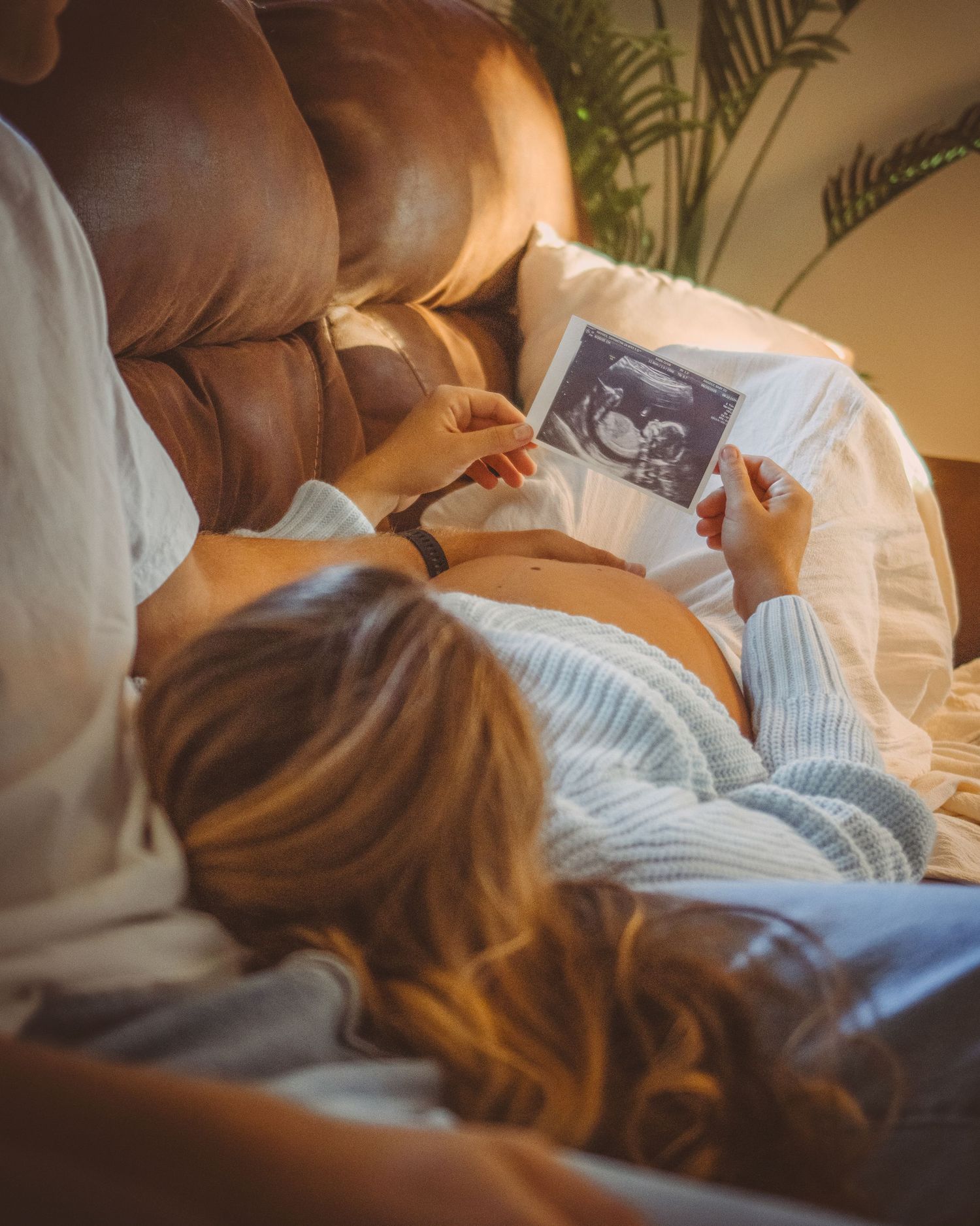 Pregnant woman lying on her husbands lap on a couch and holding ultrasound photo, showing baby bump in cozy warm setting.