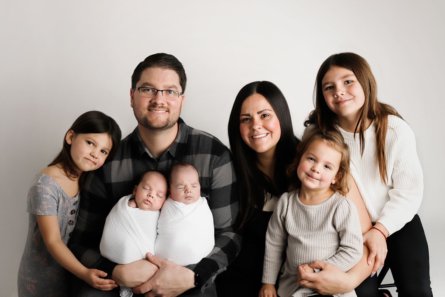Family portrait with parents holding newborn twins, flanked by three young daughters, against a white studio background. Utah photographer.