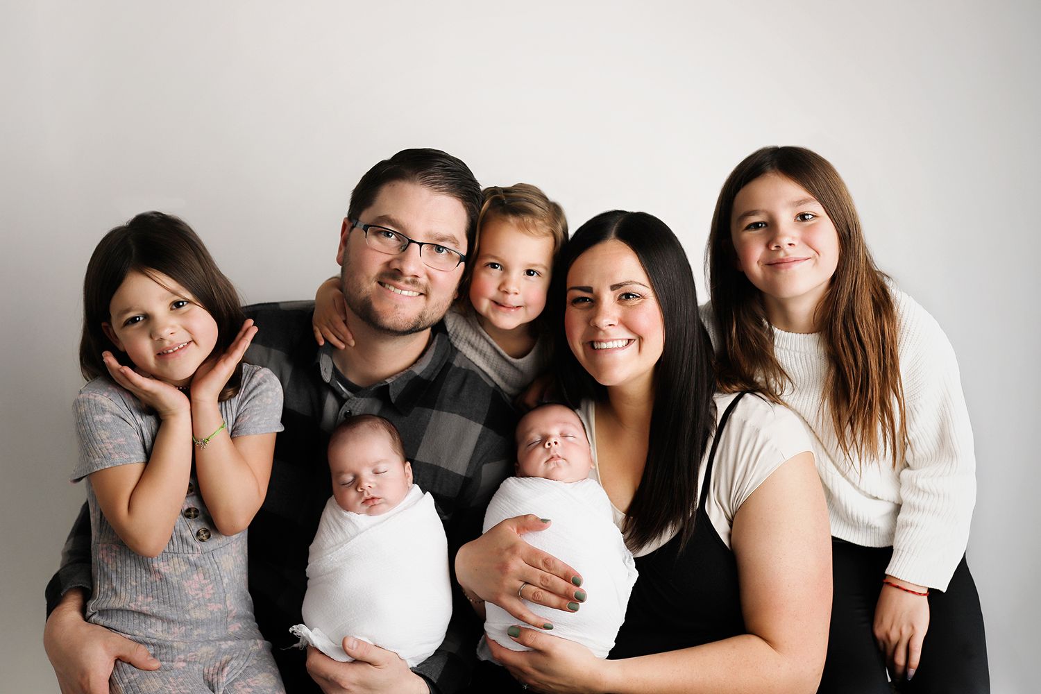 Happy family of seven posing together, including parents holding newborn twins and three older children, against a white background.