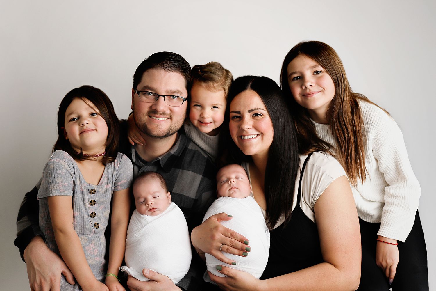 Happy family portrait with parents holding newborn twins, surrounded by three older children, against a white background. Orem family photos