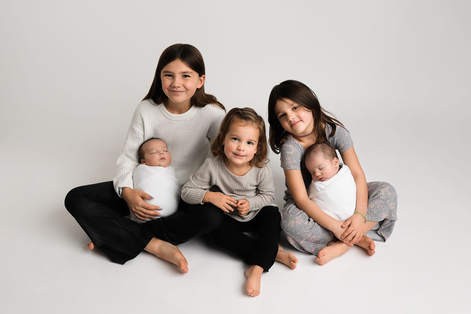 Five siblings pose together on white backdrop, two older girls holding newborn twins while a toddler sits in the middle.