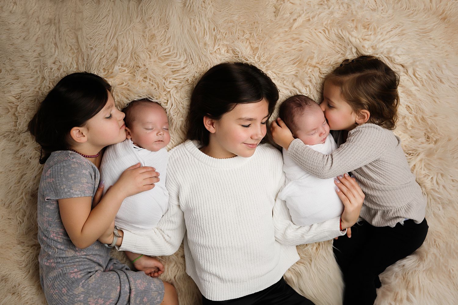 A mother lies on a fur rug with twin newborns as two older siblings lean in to give gentle kisses. Utah County twin newborn photographer.