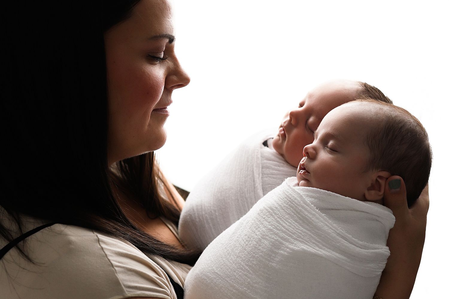 Mother tenderly holds two swaddled newborn twins wrapped in white blankets in a studio portrait.