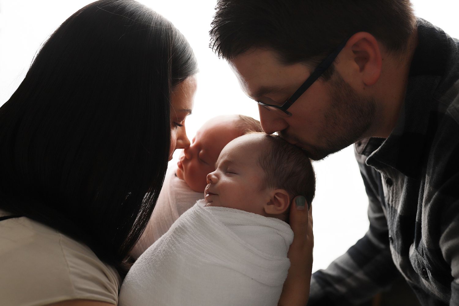Parents lovingly kissing their newborn baby wrapped in white swaddle blanket. Twin newborn photographer, Utah County photographer.