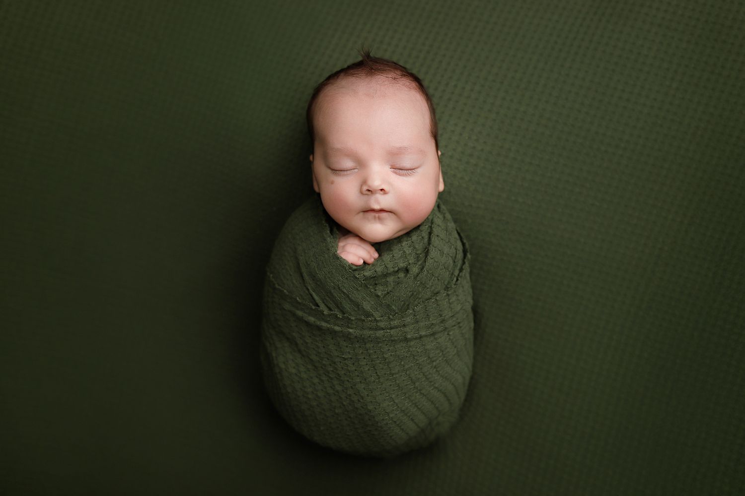 Newborn baby swaddled in green wrap sleeping peacefully on matching green textured background.