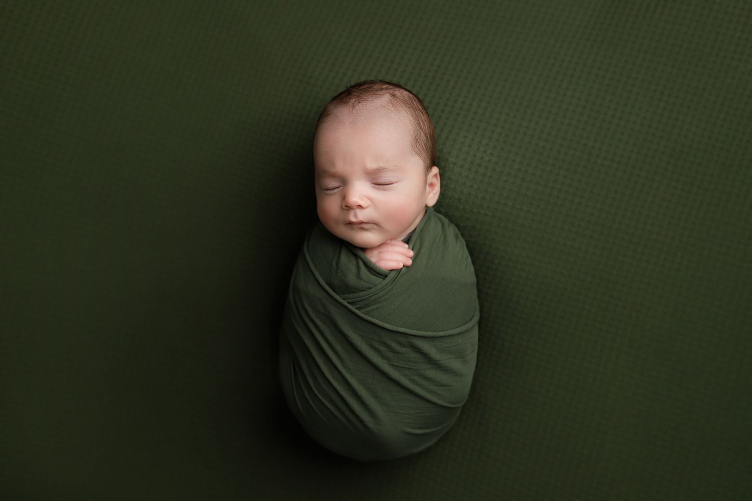 Newborn baby swaddled in green wrap sleeping peacefully on matching green textured background. Utah newborn photographer.