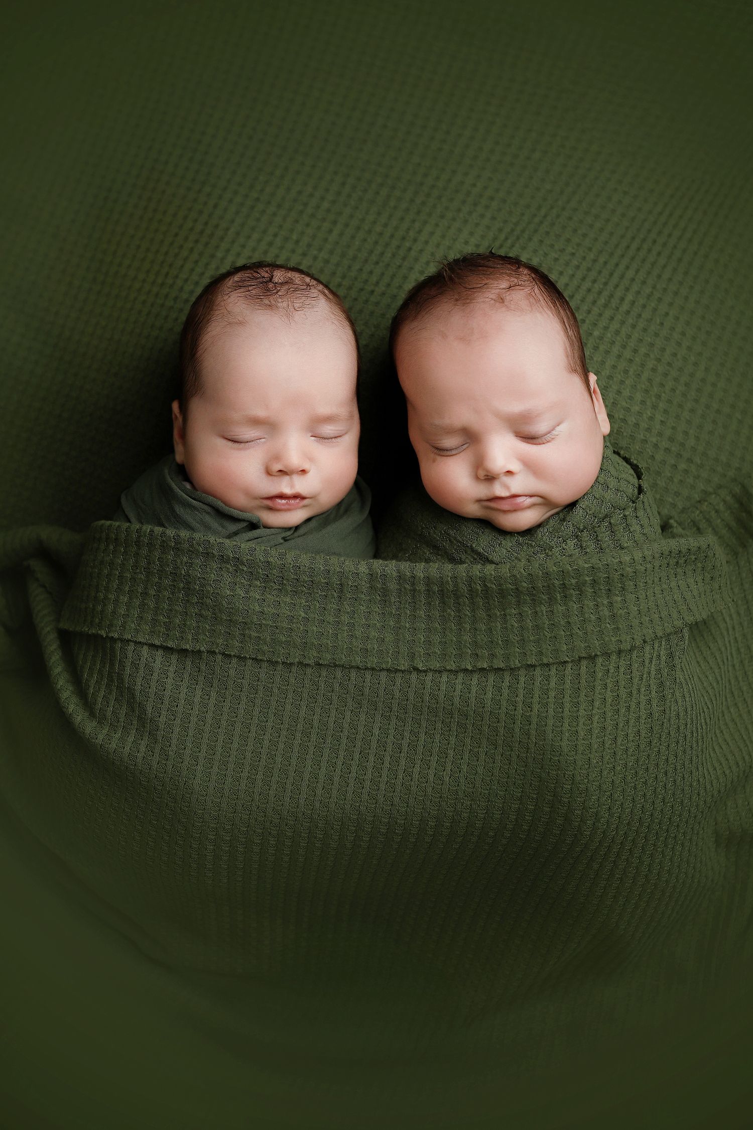 Two newborn twins swaddled in green knit wraps sleeping peacefully side by side on a green textured background. Utah County twin photography