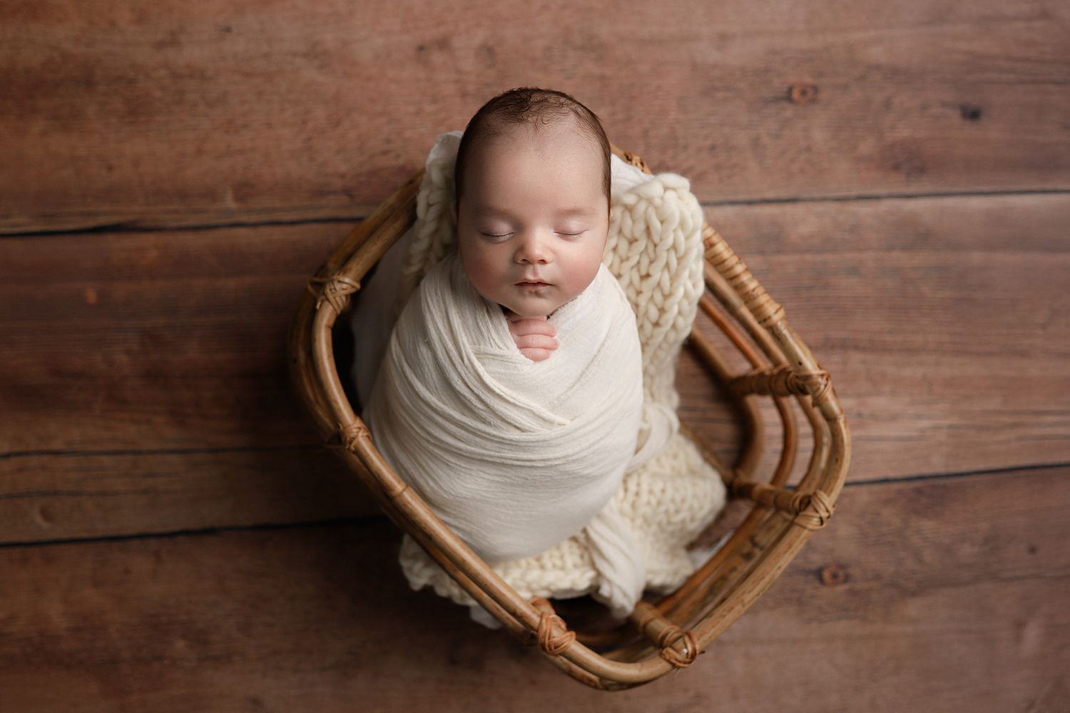 Newborn baby swaddled in cream wrap sleeping peacefully in a wicker basket on a wooden background.