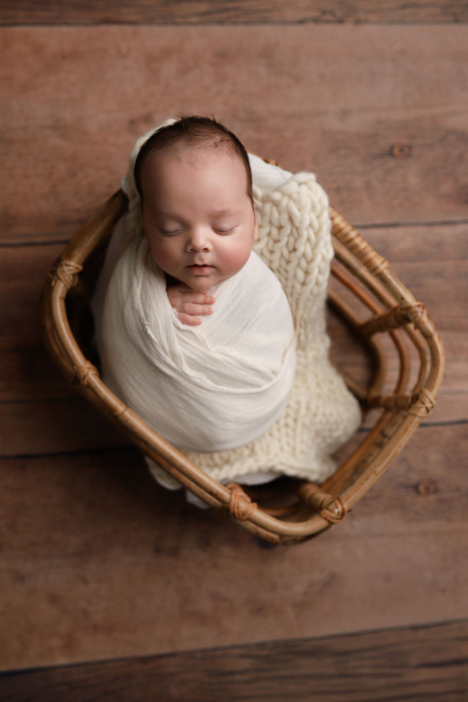 Sleeping newborn baby swaddled in white wrap inside a wicker basket with cream knit blanket on wood floor. Utah newborn photographer
