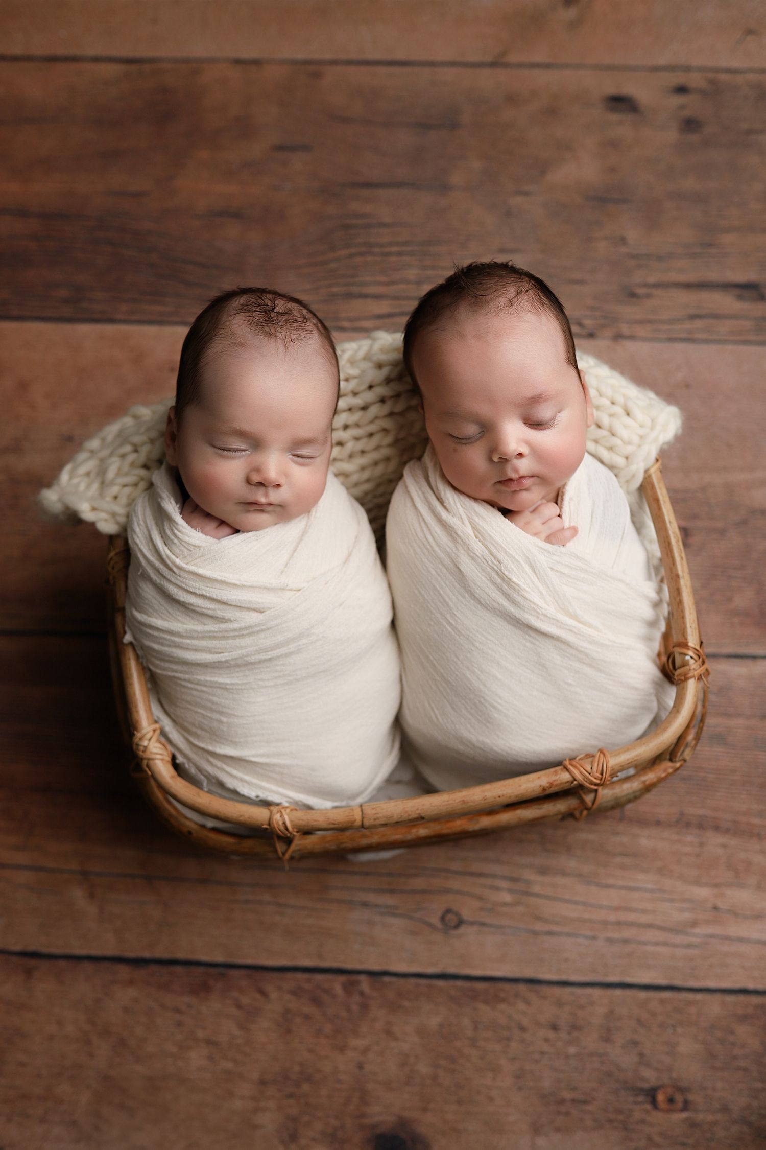 Two sleeping newborn twins swaddled in white wraps nestled together in a wicker basket on rustic wood. Utah County newborn photography