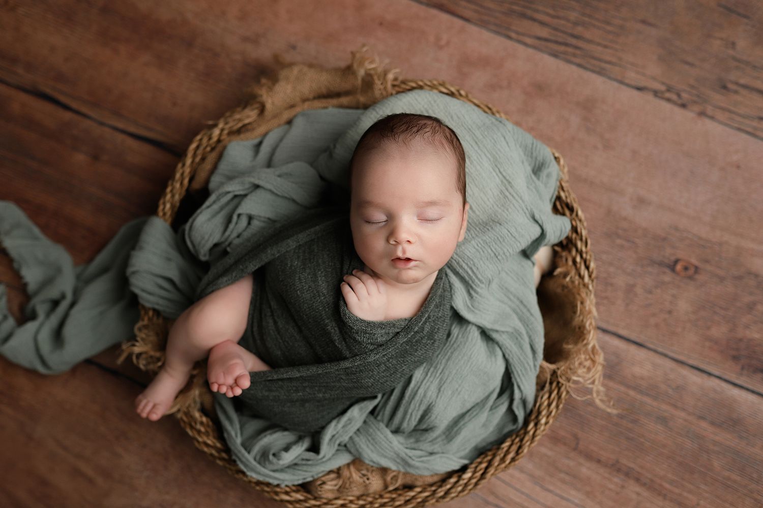 Newborn baby sleeping peacefully in a wicker basket, wrapped in a soft sage green swaddle on a wooden floor. Orem baby photographer.