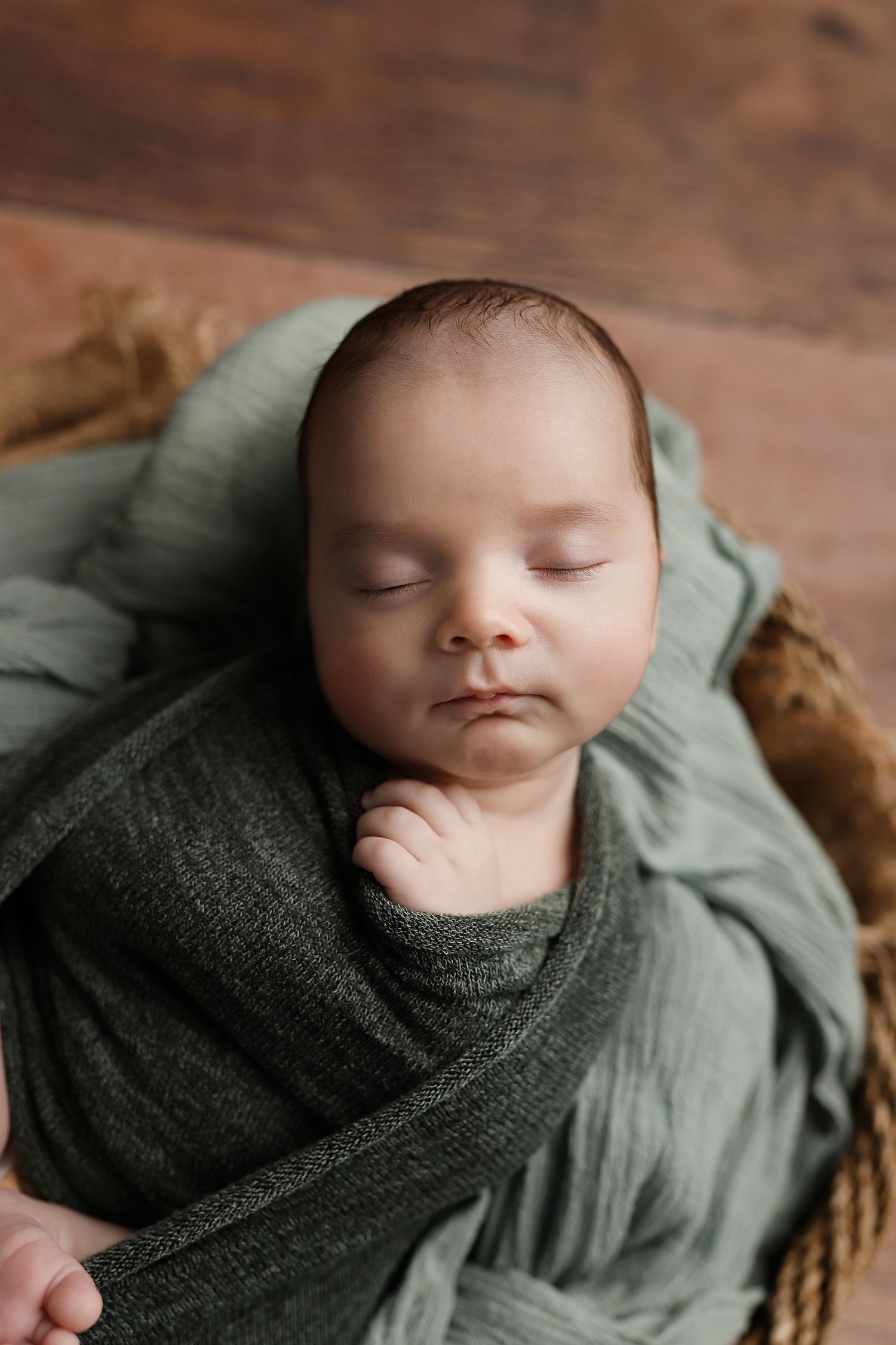 Newborn baby sleeping peacefully wrapped in a green swaddle blanket inside a wicker basket on a wooden floor. Utah newborn photographer