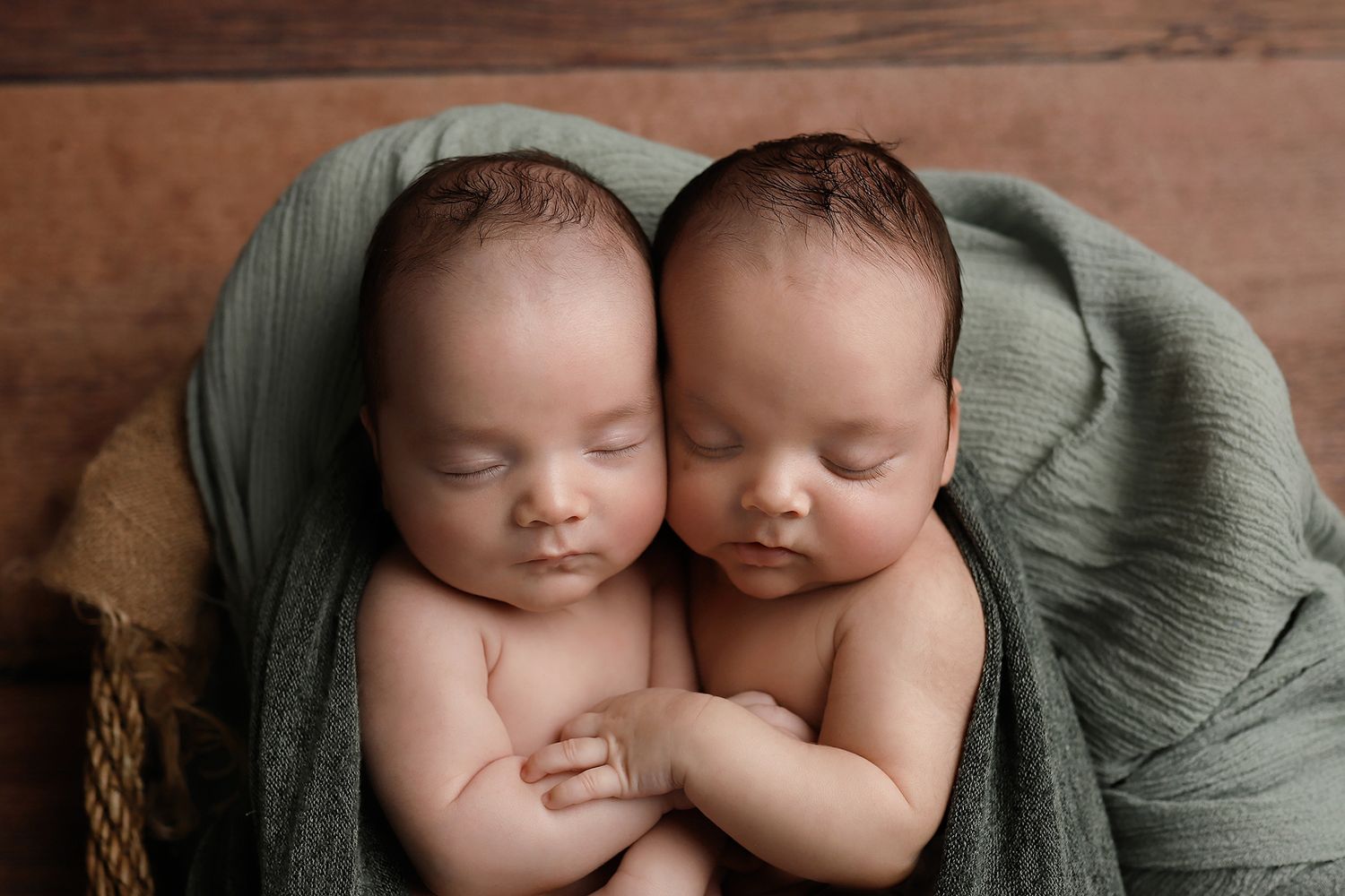 Two newborn twins sleeping peacefully wrapped in a soft green blanket inside a rustic wooden bowl. Utah County twin newborn photographer.
