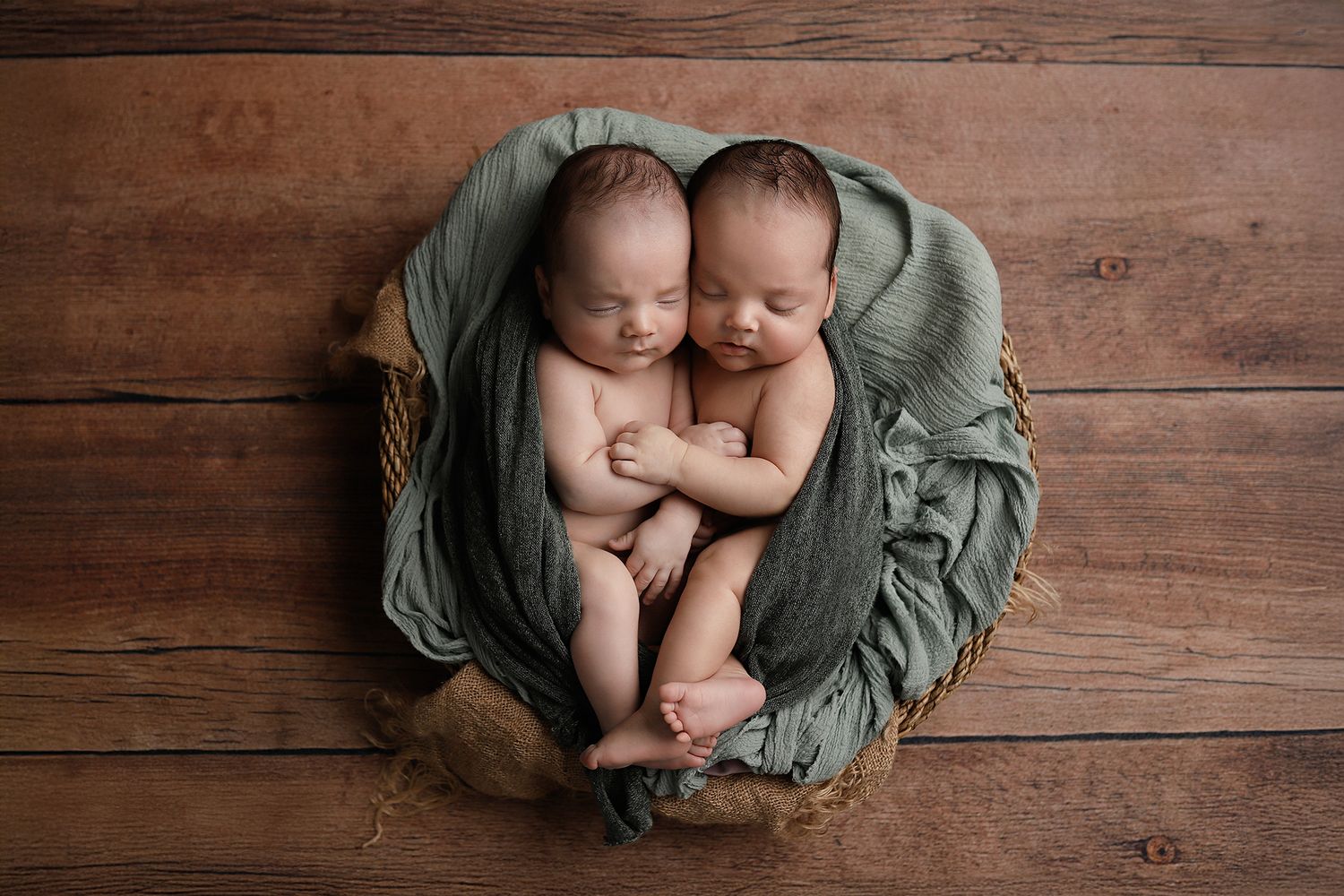 Two newborn twins sleeping peacefully wrapped in a sage green cloth inside a rustic wooden bowl on a wood backdrop. Twin newborn photos.