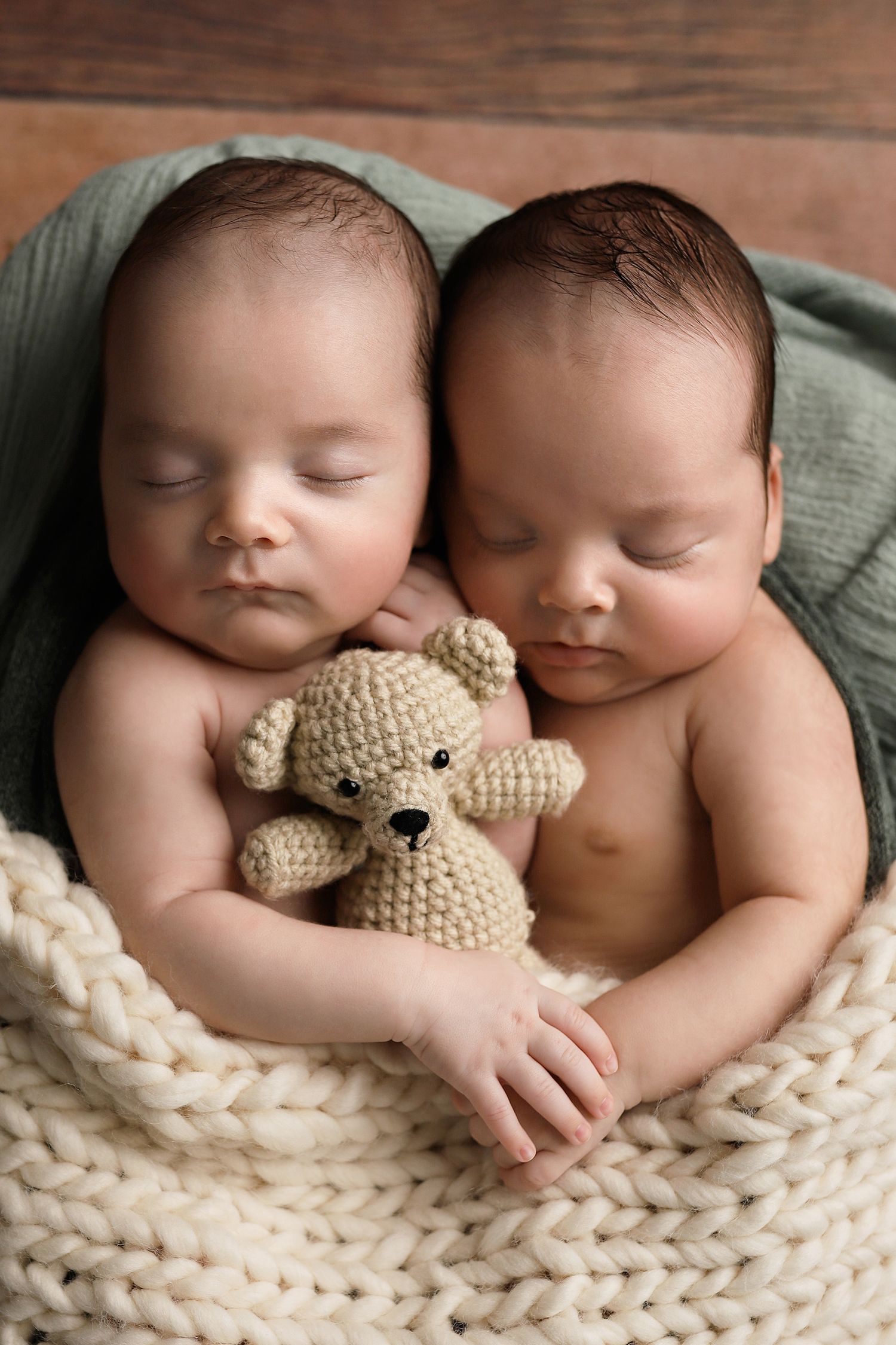 Two sleeping newborn twins cuddle together holding a small crocheted teddy bear on a soft knit blanket. Utah newborn photographer.