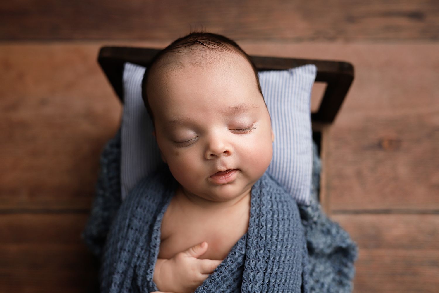 Newborn baby sleeping peacefully wrapped in a blue knit blanket in a rustic wooden bed with a striped pillow.
