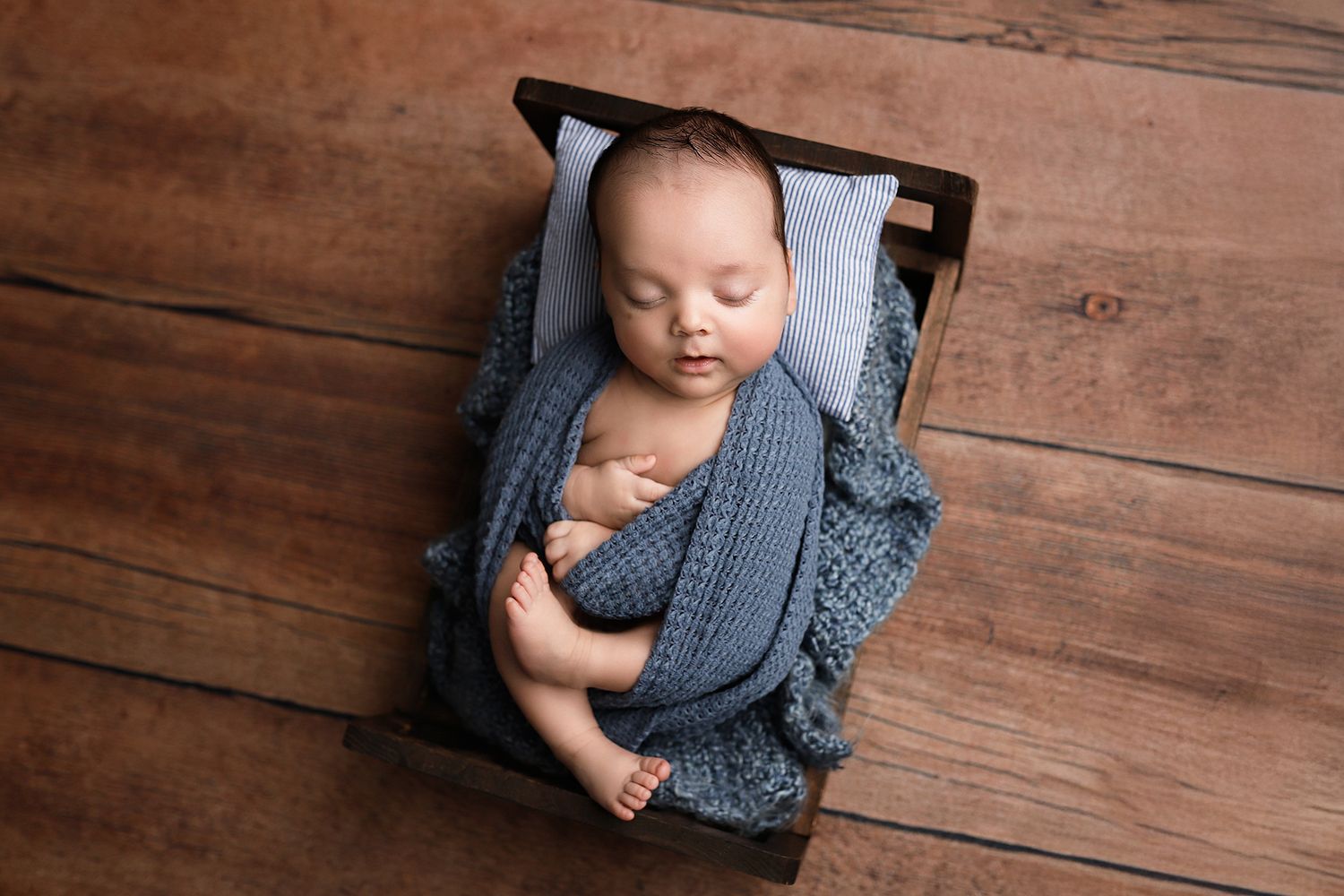 Newborn baby sleeping peacefully wrapped in blue knit blanket inside a rustic wooden crate on hardwood floor. Orem newborn photography.