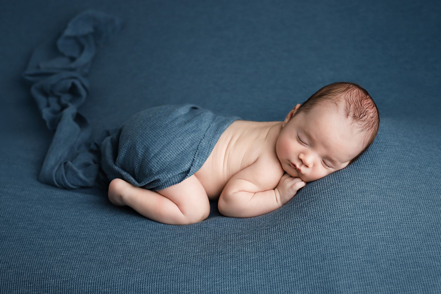 A peaceful newborn baby sleeping curled up on a soft blue blanket, wrapped in a matching blue swaddle cloth.