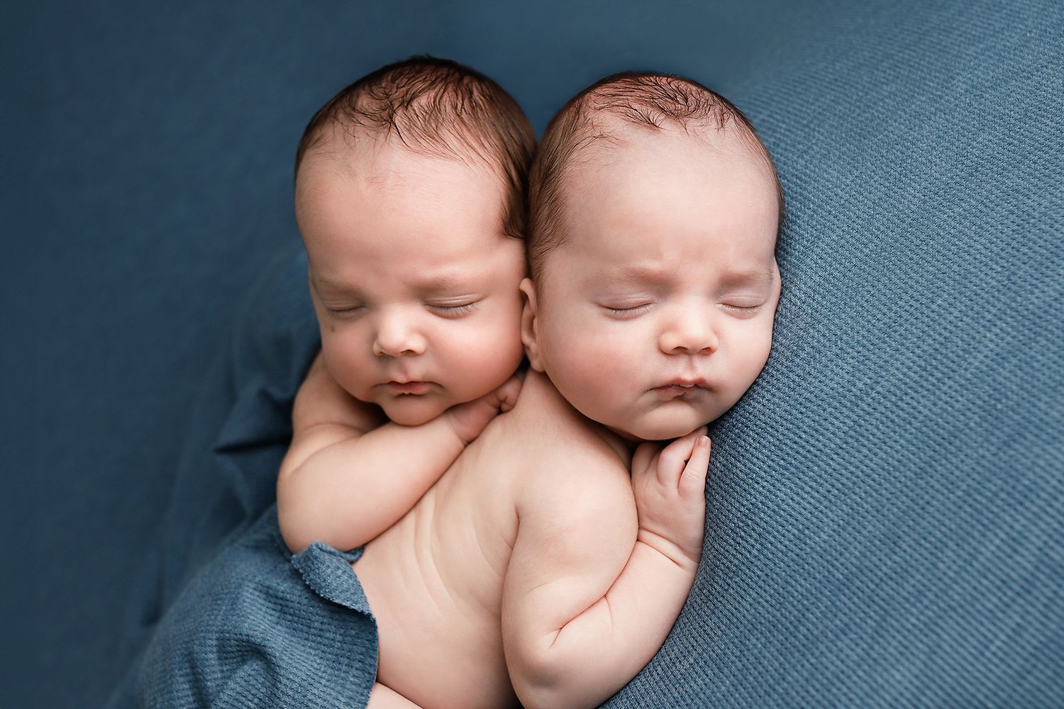 Two sleeping newborn twins cuddled together on a blue blanket in a peaceful studio newborn photography pose. Twin newborn photography.