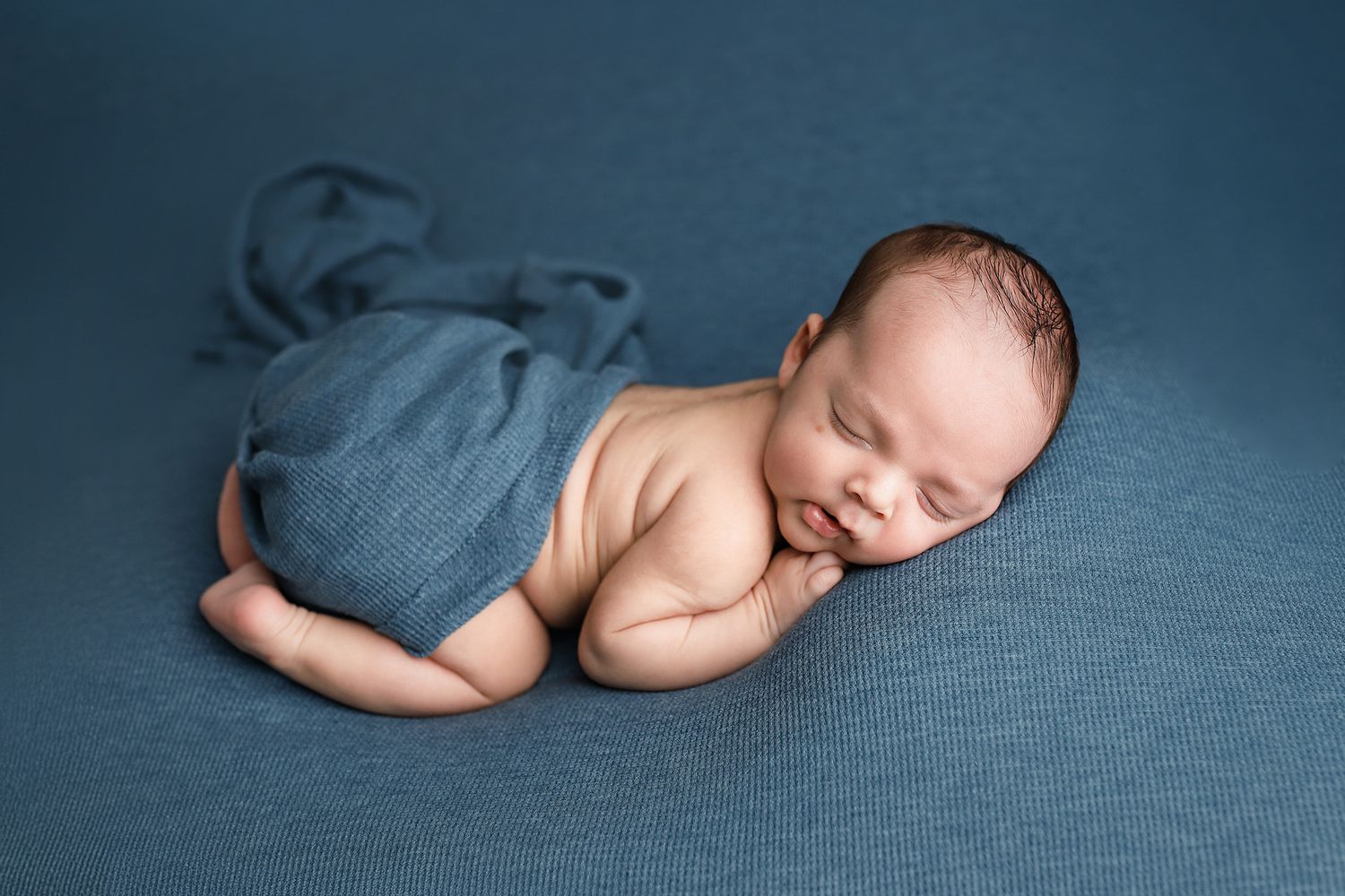 Peaceful newborn baby sleeping wrapped in a blue knit blanket on a teal backdrop in a professional photo. Utah newborn photography