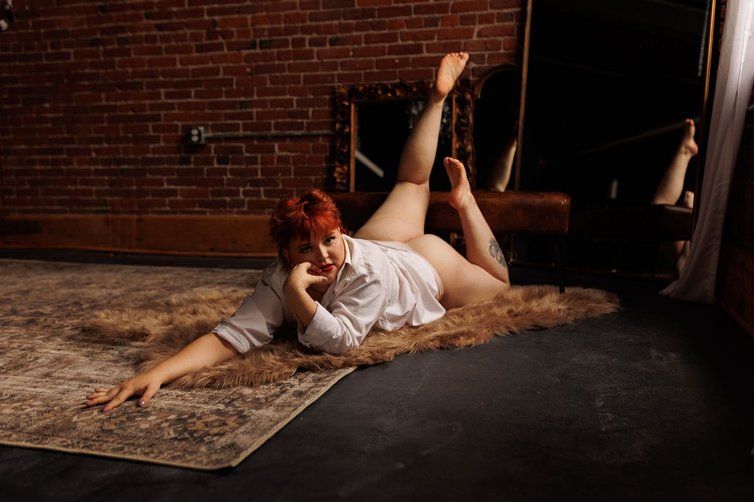 Woman with red hair poses on a vintage rug in a white shirt inside a rustic brick-walled studio.
