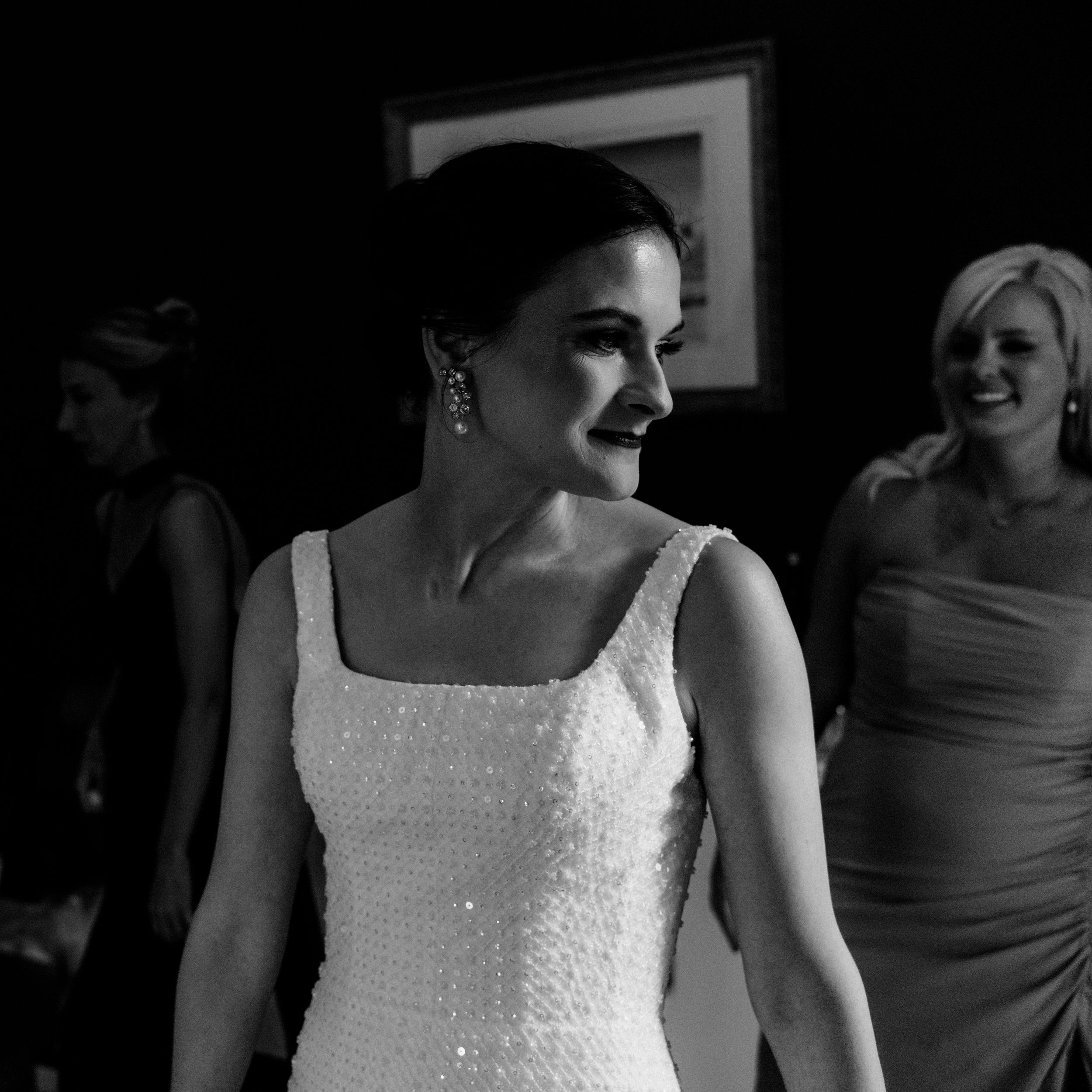 Artistic wedding photo of a bride in a sophisticated gown smiling, with bridesmaids in the background in dramatic lighting.