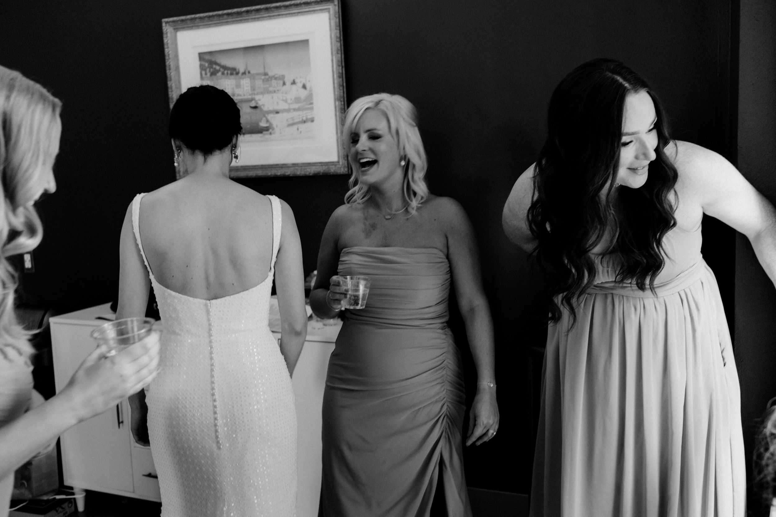 Bridesmaids laughing and holding drinks while helping bride get ready in black and white wedding photo.