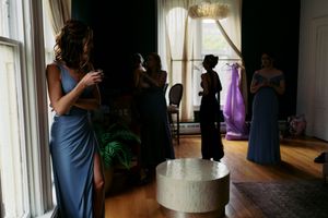 Bridesmaids in blue dresses gather in an elegant room with bride's mother silhouetted in draped window.
