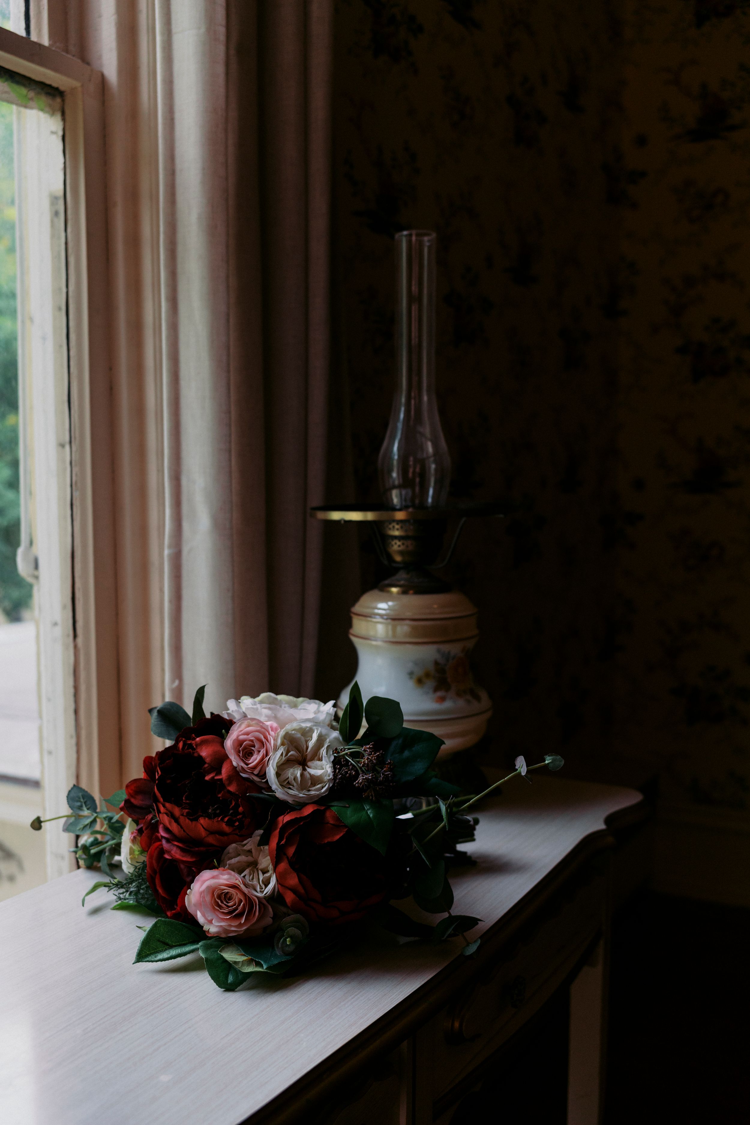 A romantic bouquet of red and pink roses beside a vintage oil lamp on a wooden table near a window.