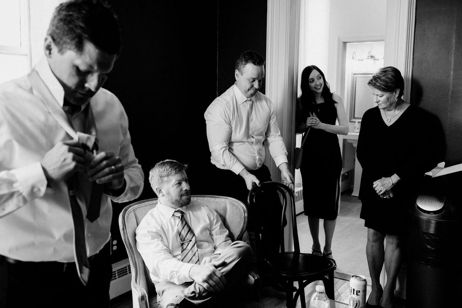 Black and white photo of a group of people socializing in groomsmen suite, with a man adjusting his tie in layered, photojournalistic style.