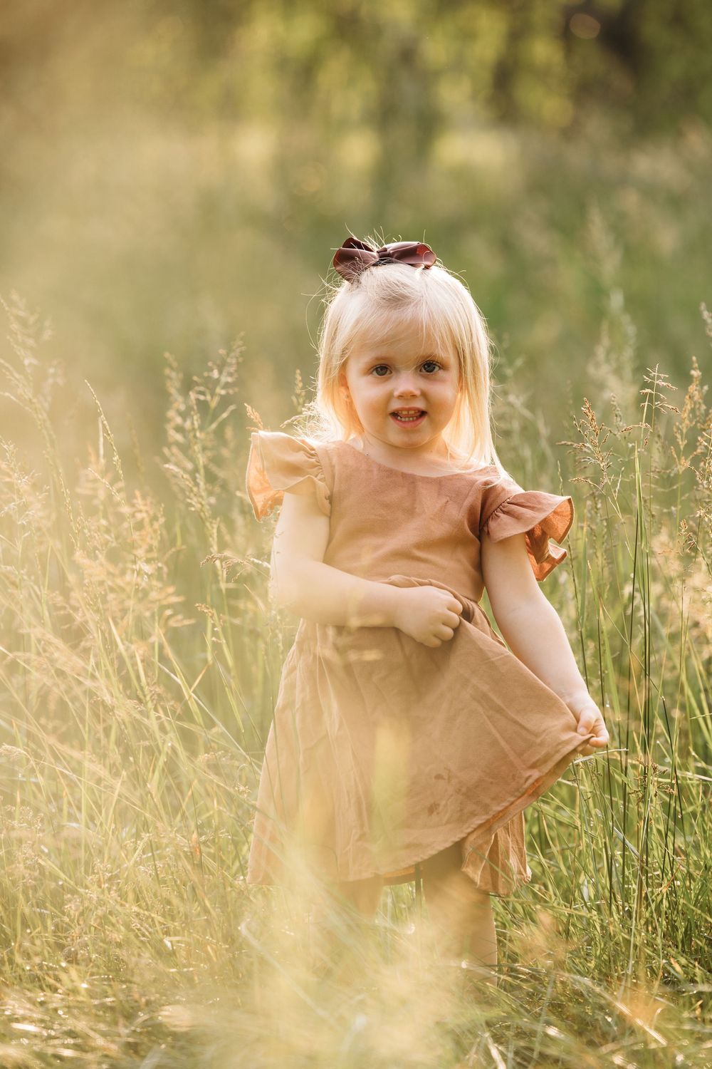 Smiling blonde toddler girl in a dusty rose dress standing in a sunlit meadow with tall golden grass.