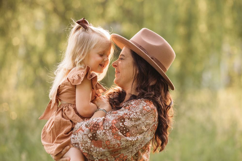 A mother in a tan hat holds her young blonde daughter close as they smile at each other outdoors in golden sunlight.