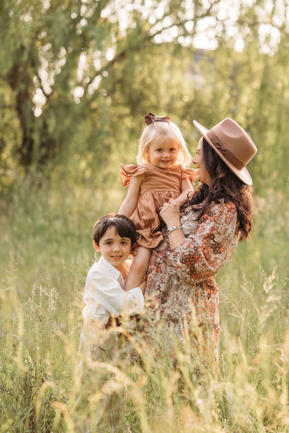 A mother in a floral dress and hat holds her toddler daughter while her young son smiles in a sunny meadow.