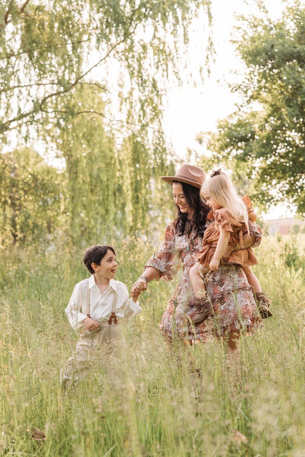 A joyful mother in a floral dress and hat holds a toddler while a young boy laughs in a sunlit meadow.
