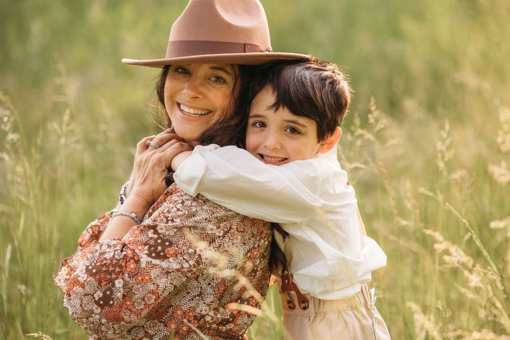 A smiling woman in a floral dress and tan hat hugs a young child in a sunny green meadow.