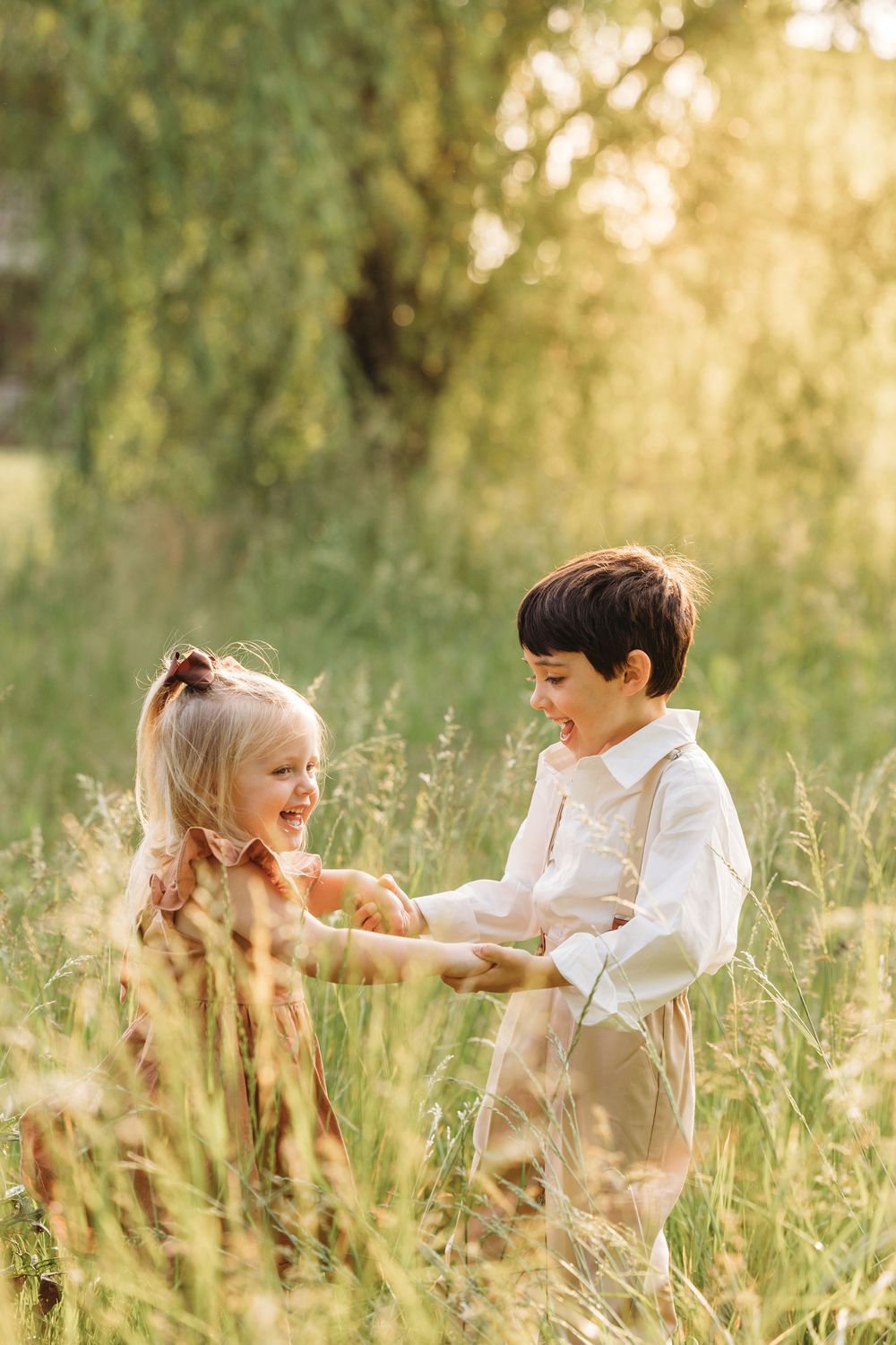 Two children holding hands and laughing together in a sunlit meadow with tall grass and trees in the background.