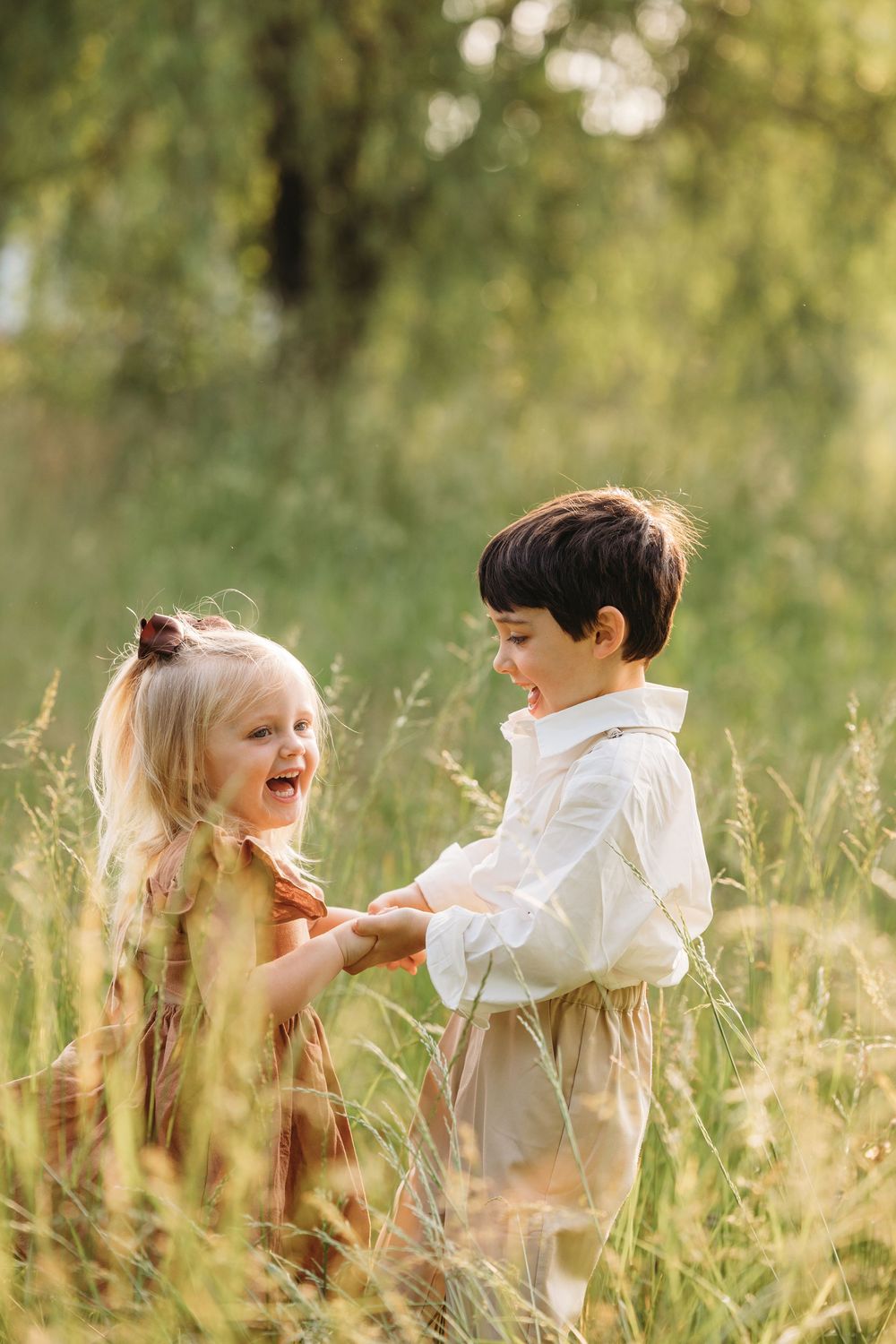 Two happy children holding hands in a sunlit meadow, a blonde girl in a rust dress and a boy in a white shirt smiling.
