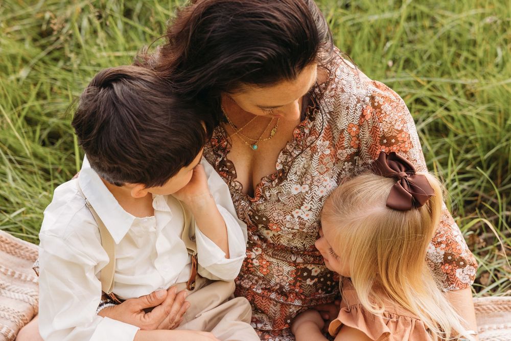 A mother in a floral dress sits on a blanket in a field, tenderly embracing her young son and toddler daughter.