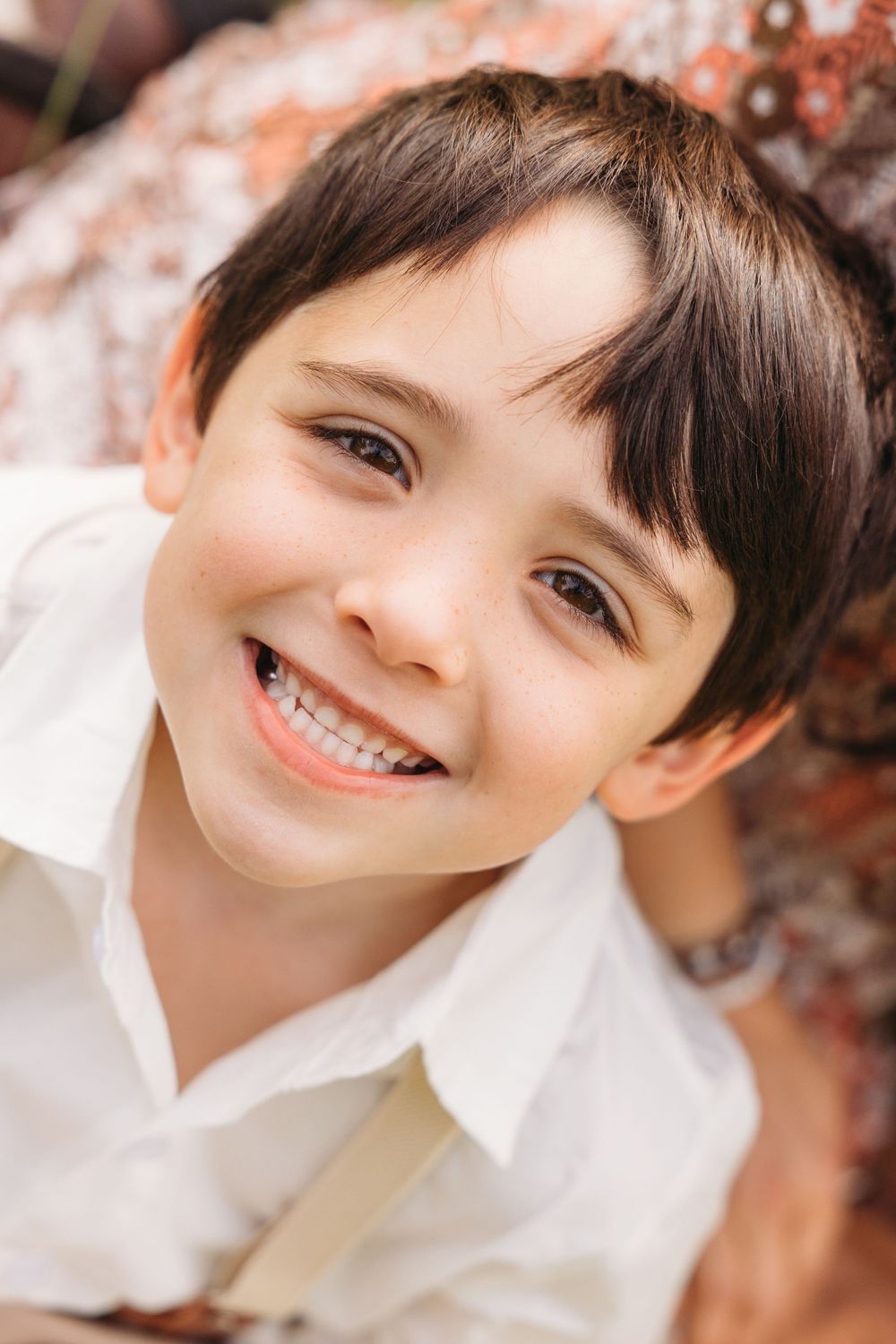 Smiling young boy with dark hair wearing a white shirt, looking up at the camera with a cheerful expression outdoors.
