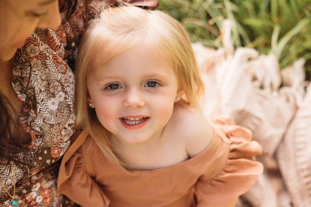 Smiling toddler girl with blonde hair wearing a rust orange dress, sitting outdoors in a candid portrait photo.