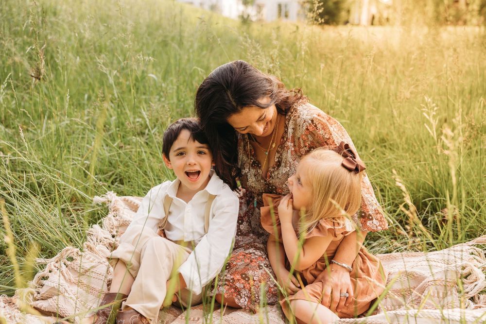 A mother sits in a grassy field with two young children, sharing a joyful moment on a blanket outdoors.