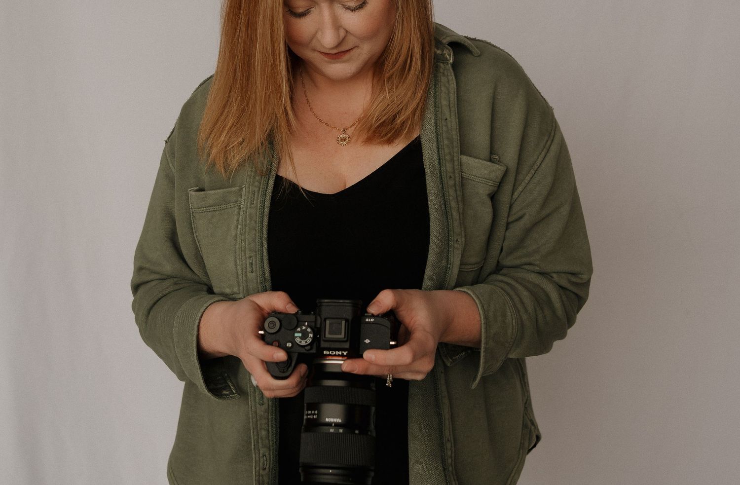 Woman in green jacket holding a camera, looking down at the screen in a photography studio setting.