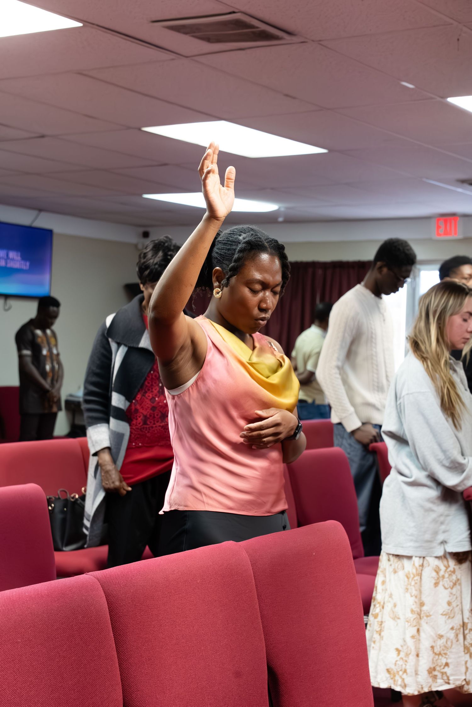 Woman raising hand in worship at church service with congregation members standing among red pews.
