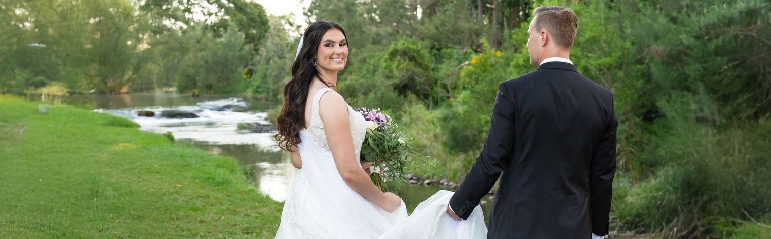 Bride and groom walking hand in hand along a lush green riverside on their wedding day.