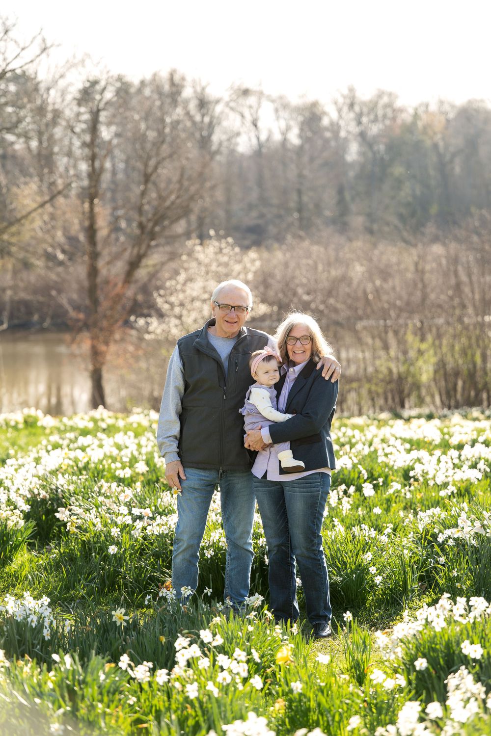 Grandparents stand smiling with their baby grandchild in a sunny field of white daffodils near a pond.