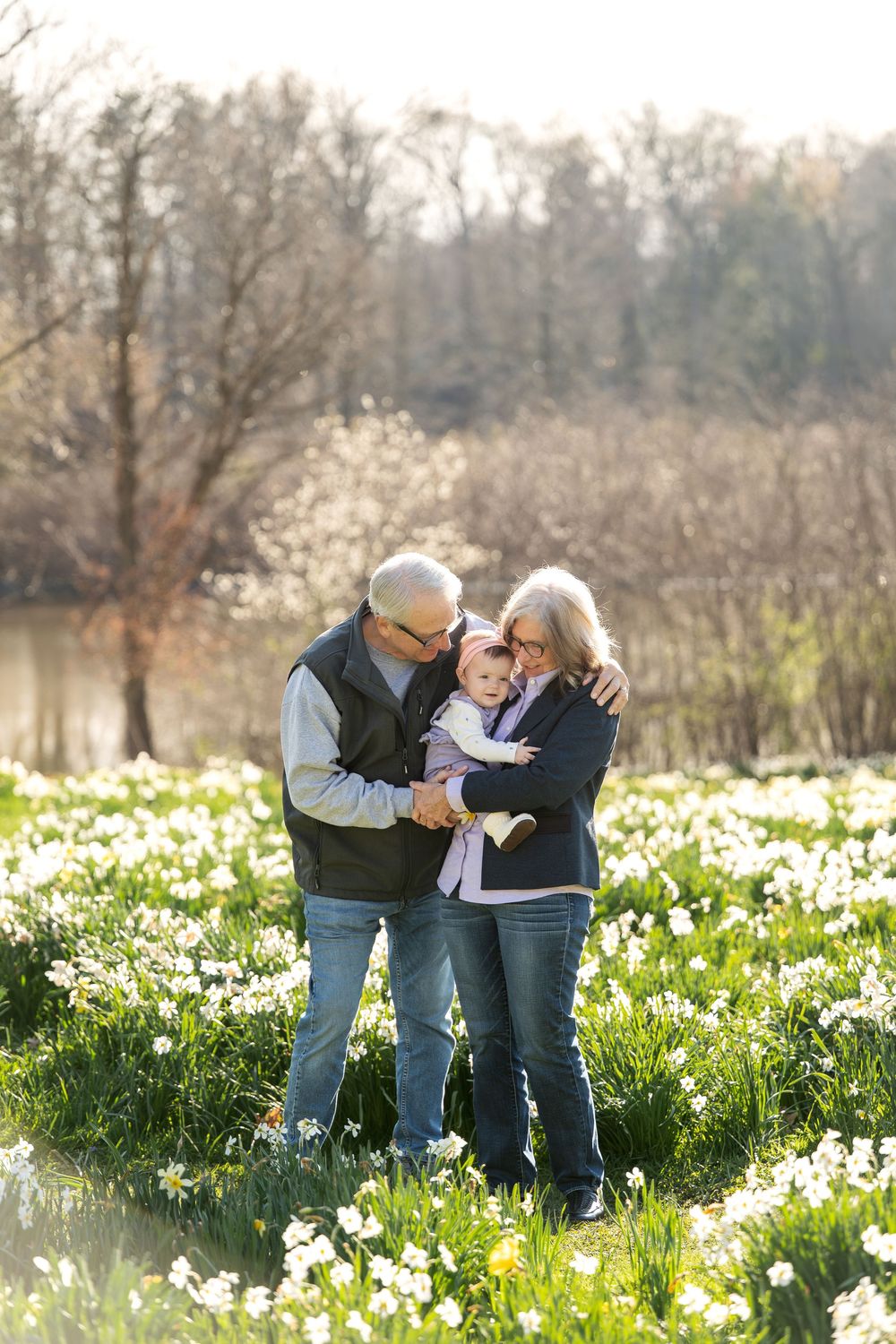 Grandparents cuddle and kiss their baby grandchild while standing in a blooming spring daffodil field.