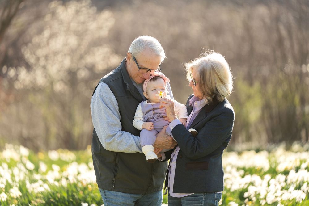 Grandparents lovingly hold their infant grandchild in a sunlit field of white daffodil flowers.