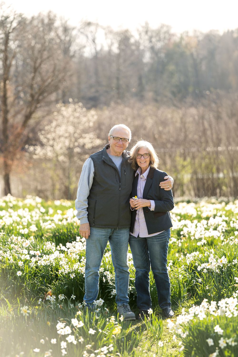 A smiling older couple embraces while standing together in a sunlit field of white daffodils in spring.